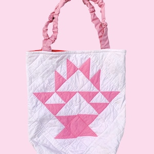 May include: A white quilted tote bag with pink patchwork in the shape of a basket. The bag has pink fabric handles.