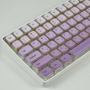 Puede incluir: Un teclado mecánico blanco con teclas moradas y blancas. Las teclas están etiquetadas con letras, números y símbolos. El teclado tiene un marco blanco y una barra espaciadora morada.