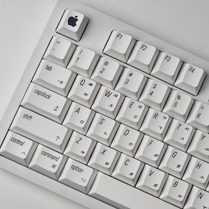 May include: A white Apple keyboard with a silver Apple logo on the top left key. The keyboard has a standard layout with function keys at the top, a number row, and letter keys below. The keys are white with black lettering.