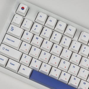 Könnte beinhalten: Weiße Tastatur mit blauen und roten Tastenkappen. Die Tastatur hat ein koreanisches Tastaturlayout mit englischen Buchstaben und koreanischen Zeichen auf den Tasten. Die Tastatur hat eine blaue Leertaste.