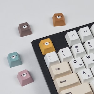 May include: Five keycaps with bear faces, in different colors, including blue, pink, brown, yellow, and orange. The keycaps are on a white surface, with a black keyboard in the background. The keyboard has keys labeled with letters, numbers, and symbols.