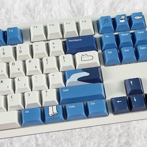 May include: A white mechanical keyboard with blue keycaps. The keycaps feature various symbols and designs, including a cat, a wave, and footprints. The keyboard has a white frame and a white background.