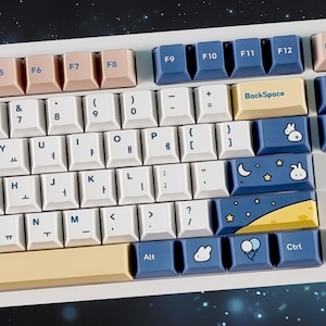 May include: A white keyboard with blue and yellow keycaps featuring a moon, stars, and a bunny rabbit design. The keyboard has a space bar with a yellow and blue design and the words "BackSpace", "Alt", and "Ctrl" on the keys.