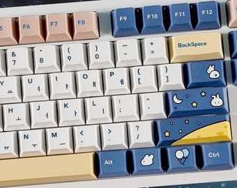 May include: A white keyboard with blue and yellow keycaps featuring a moon, stars, and a bunny rabbit design. The keyboard has a space bar with a yellow and blue design and the words "BackSpace", "Alt", and "Ctrl" on the keys.