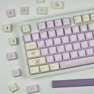 Könnte beinhalten: Eine weiße Tastatur mit lila und weißen Tasten. Die Tasten haben verschiedene Symbole und Buchstaben darauf gedruckt. Die Tastatur hat eine lila und weiße Leertaste und eine lila und weiße Handballenauflage. Die Tasten haben niedliche Illustrationen von Katzen und anderen Designs.