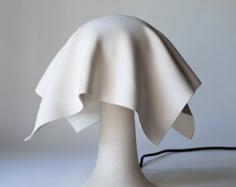 May include: A white ceramic table lamp with a fabric shade. The shade is draped over the top of the lamp and has a soft, flowing texture.