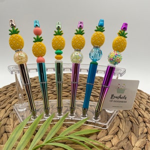 May include: Six colorful pineapple-themed pens with iridescent accents. The pens are displayed in a clear plastic holder on a woven surface. The text "Bushels of Goodness" is visible on a white tag.
