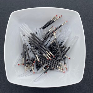 May include: A white ceramic bowl filled with a large quantity of black and silver ballpoint pen refills with red tips.