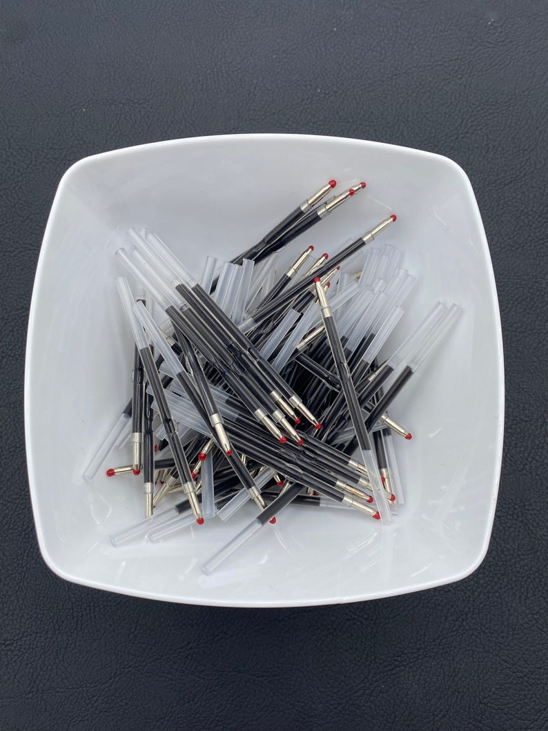 May include: A white ceramic bowl filled with a large quantity of black and silver ballpoint pen refills with red tips.