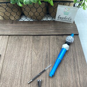 May include: A blue and white volleyball pen with a black and white volleyball charm. The pen is lying on a wooden surface. The text "bushels of goodness" is visible on a sticker in the background.