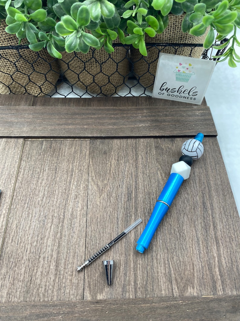 May include: A blue and white volleyball pen with a black and white volleyball charm. The pen is lying on a wooden surface. The text "bushels of goodness" is visible on a sticker in the background.