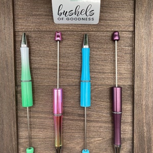 May include: Four pens with different colored barrels and silver accents. The pens are displayed on a wooden background. The pens are green, pink, blue, and purple.