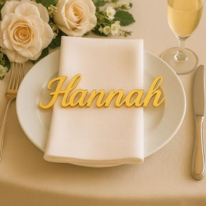 May include: A gold-colored nameplate that reads "Hannah" rests on a white napkin atop a white plate. A fork and knife are on either side of the plate. A glass of champagne and white roses are in the background. The table setting is elegant.