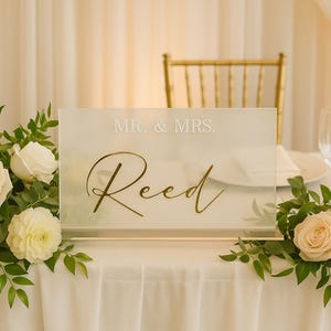 May include: A clear acrylic wedding sign with the text "MR. & MRS. Reed" in gold script. The sign is displayed on a white tablecloth, surrounded by floral arrangements of white and peach roses and greenery.
