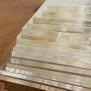 Puede incluir: Una pila de posavasos rectangulares transparentes de acr&iacute;lico. Los posavasos est&aacute;n dispuestos en un patr&oacute;n escalonado, con cada capa ligeramente desplazada. Los bordes de los posavasos tienen una apariencia sutil y texturizada.