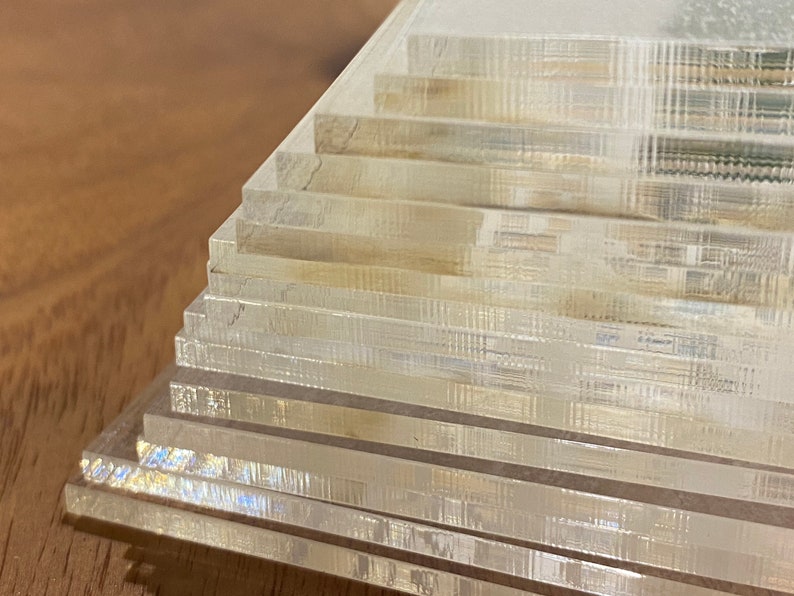 Puede incluir: Una pila de posavasos rectangulares transparentes de acr&iacute;lico. Los posavasos est&aacute;n dispuestos en un patr&oacute;n escalonado, con cada capa ligeramente desplazada. Los bordes de los posavasos tienen una apariencia sutil y texturizada.