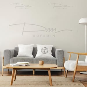 May include: A gray sofa with two white pillows, one with a black and white graphic design. A wooden coffee table with a book and a cup on it. A wooden chair with a white cushion. The wall behind the furniture is light gray with the text "Dofamin" written in gray.