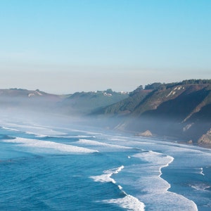 Peut inclure: Paysage côtier avec une vaste étendue d'océan et des vagues déferlant sur une plage de sable. L'image montre un ciel bleu clair, avec des collines et des falaises le long de la côte, partiellement voilées par une légère brume.
