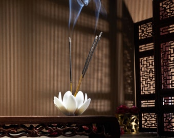 May include: A white lotus flower-shaped incense holder with three burning incense sticks, emitting wisps of smoke. The holder sits on a dark wooden stand with ornate carvings. The background features a brown textured wall and a decorative screen.