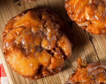 May include: Close-up of several glazed apple fritters on a wooden cutting board. The fritters are golden brown with a glossy, white glaze. A red and white checkered cloth is visible in the background, suggesting a rustic setting.