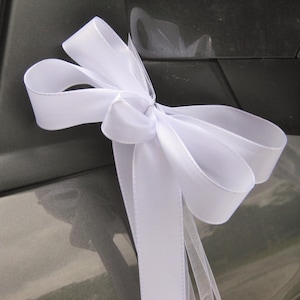 May include: A white satin ribbon bow, likely for a wedding or celebration, is attached to a dark gray car door. The bow is large and has multiple loops and streamers, creating a decorative accent.