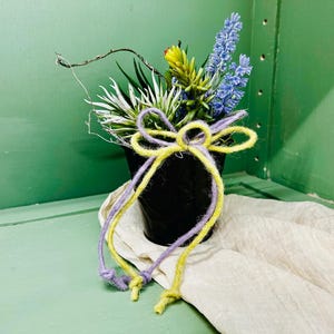 Op de afbeelding: Een kleine zwarte pot gevuld met kunstbloemen in blauw, geel en wit. Een decoratieve strik van geel en paars garen is om de pot gebonden. De opstelling staat op een witte doek en creëert een kleurrijk display.