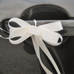 Puede incluir: Un lazo de cinta de satén blanco atado a la manija de la puerta de un coche. El lazo está hecho de cinta ancha y lisa con largas colas que fluyen. La manija de la puerta del coche es de color gris oscuro.