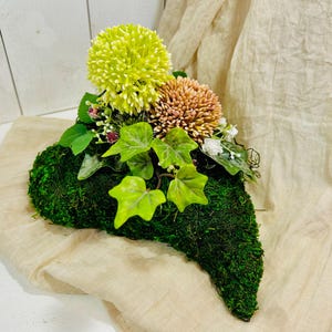Könnte beinhalten: Ein herzförmiger Moosuntergrund trägt ein Blumenarrangement. Das Arrangement zeigt eine große, chartreusefarbene Pomponblume, eine kleinere pfirsichfarbene Blume sowie Zweige mit weißen Blüten und Grün. Grüne Efeublätter sorgen für einen natürlichen Touch.