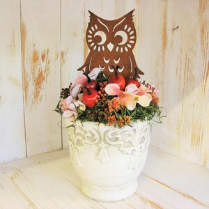 Può includere: Un vaso di ceramica bianco e ornato pieno di una composizione floreale, tra cui mele rosse, fiori rosa e arancioni e verde. Una silhouette di gufo in metallo marrone si trova sopra la composizione. Lo sfondo è un muro di legno bianco.
