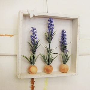 Puede incluir: Una caja de sombra de madera blanca y cuadrada con tres ramitas de lavanda. Cada ramita tiene flores moradas, hojas verdes y una base color melocotón. Una mariposa blanca decora la parte superior de la caja. La caja está montada en una pared blanca.