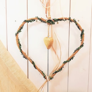 Könnte beinhalten: Ein herzförmiger Kranz aus Schnur und Grün hängt an einer weißen Holzwand. Ein kleines goldenes Herz-Ornament hängt am Kranz.