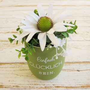 Può includere: Un piccolo vaso di ceramica verde con la scritta bianca "Einfach Glücklich Sein", riempito con un fiore bianco a forma di margherita e fogliame verde. Il fiore ha un centro verde e marrone. Il vaso è su una superficie di legno bianco.
