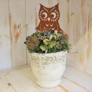 Può includere: Un vaso di ceramica bianco e ornato, pieno di fiori secchi e verde, sormontato da una sagoma di gufo in metallo marrone arrugginito. Il vaso è posto su una superficie di legno bianco contro uno sfondo di assi di legno bianche.