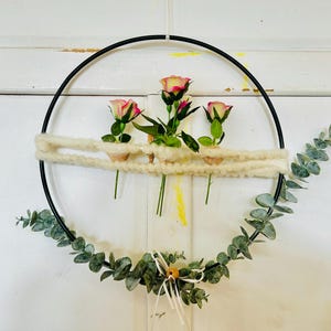Könnte beinhalten: Ein schwarzer Metall-Blumenreifen mit drei rosa-gelben Rosen, einem cremefarbenen Wollakzent und grünen Eukalyptusblättern. Das Blumenarrangement ist ein dekoratives Element für die Inneneinrichtung.