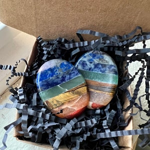 May include: Two oval-shaped, layered gemstone stones in a cardboard box with black paper filler. The stones feature bands of blue, green, yellow, orange, and brown. The box is open, revealing the stones nestled within.