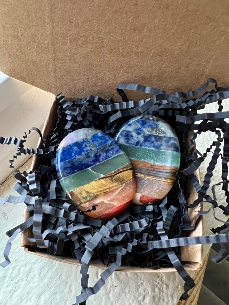 May include: Two oval-shaped, layered gemstone stones in a cardboard box with black paper filler. The stones feature bands of blue, green, yellow, orange, and brown. The box is open, revealing the stones nestled within.