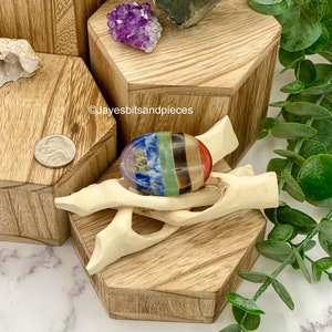 May include: A colorful chakra stone, featuring bands of red, orange, yellow, green, blue, indigo, and violet, rests on a carved wooden stand. The stone is displayed on a wooden hexagonal block, with other crystals and a nickel coin nearby.