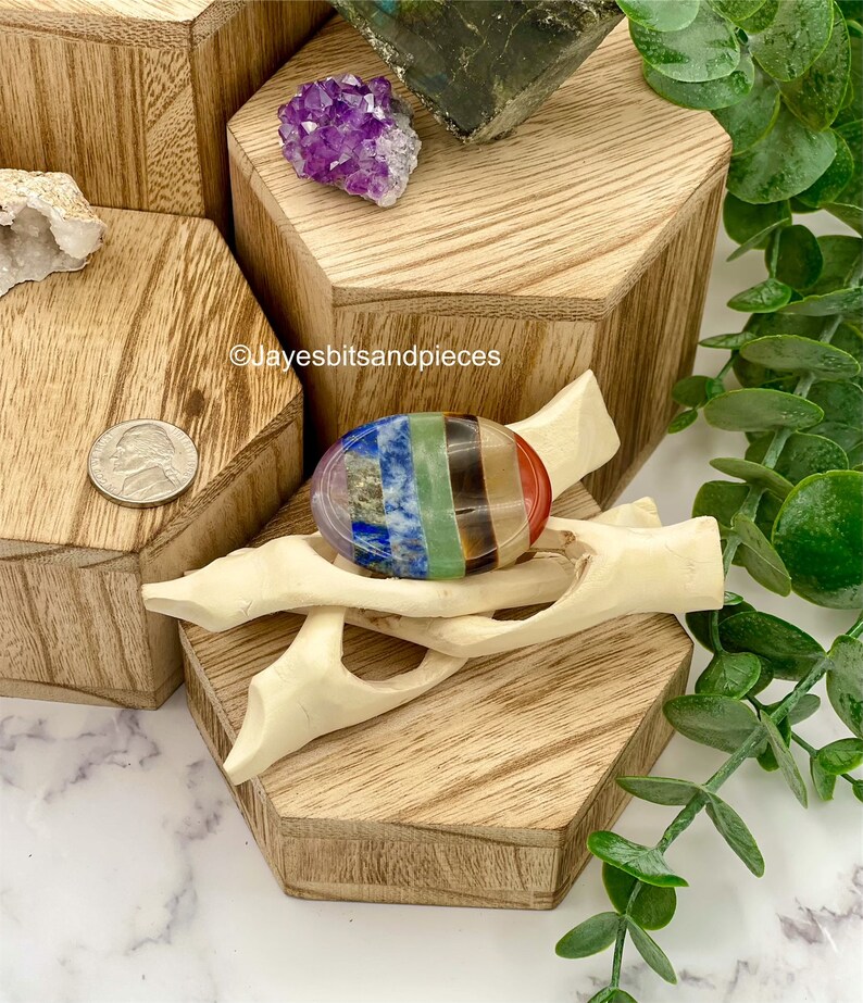 May include: A colorful chakra stone, featuring bands of red, orange, yellow, green, blue, indigo, and violet, rests on a carved wooden stand. The stone is displayed on a wooden hexagonal block, with other crystals and a nickel coin nearby.