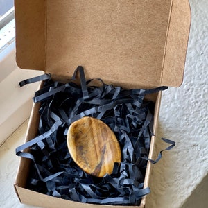 May include: A polished, oval-shaped tiger's eye stone rests on black paper shreds inside a small, open cardboard box. The stone displays a mix of golden-brown and black colors. The box is sitting on a white surface.