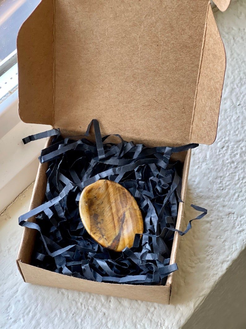 May include: A polished, oval-shaped tiger's eye stone rests on black paper shreds inside a small, open cardboard box. The stone displays a mix of golden-brown and black colors. The box is sitting on a white surface.