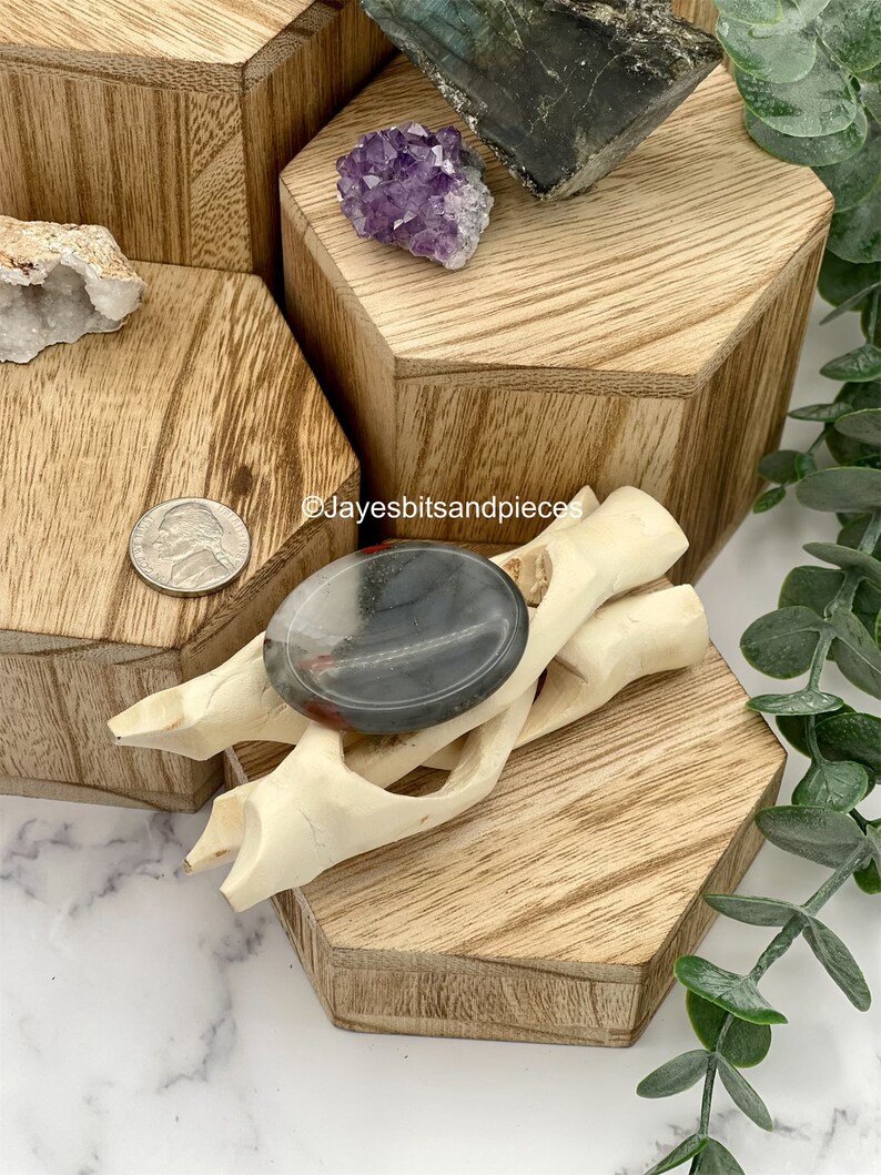 May include: A polished, oval-shaped stone with gray and red hues rests on a carved, off-white wooden accent piece. The stone is displayed on a wooden hexagonal riser, alongside other crystals and a US quarter. The background features additional wooden risers and greenery.