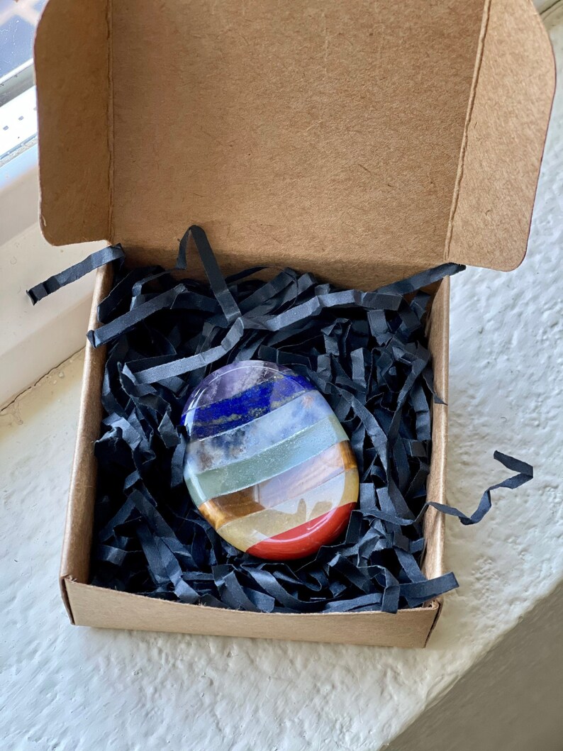 May include: A colorful, oval-shaped chakra stone is nestled in a small, open cardboard box filled with black paper strips. The stone features bands of red, orange, yellow, green, blue, and purple.