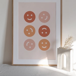 May include: A framed print featuring a pink background and six different coloured smiley faces. The smiley faces are arranged in a grid pattern with three rows and two columns. The frame is a light wood colour.