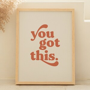 K&ouml;nnte beinhalten: Ein gerahmter Druck mit dem Text "you got this" in einem Retro-Schriftart-Stil. Der Text ist in einem verbrannten Orangeton gehalten und der Hintergrund ist hellbeige. Der Rahmen ist in einer hellen Holzfarbe.