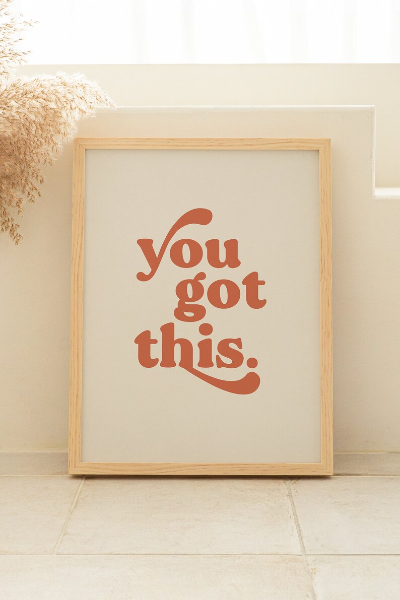 K&ouml;nnte beinhalten: Ein gerahmter Druck mit dem Text "you got this" in einem Retro-Schriftart-Stil. Der Text ist in einem verbrannten Orangeton gehalten und der Hintergrund ist hellbeige. Der Rahmen ist in einer hellen Holzfarbe.