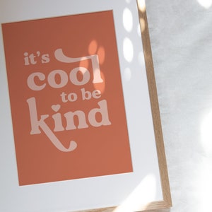 May include: Framed art print with the message "it's cool to be kind" in a retro font. The print has a terracotta background and is framed in a light wood frame with a white mat.