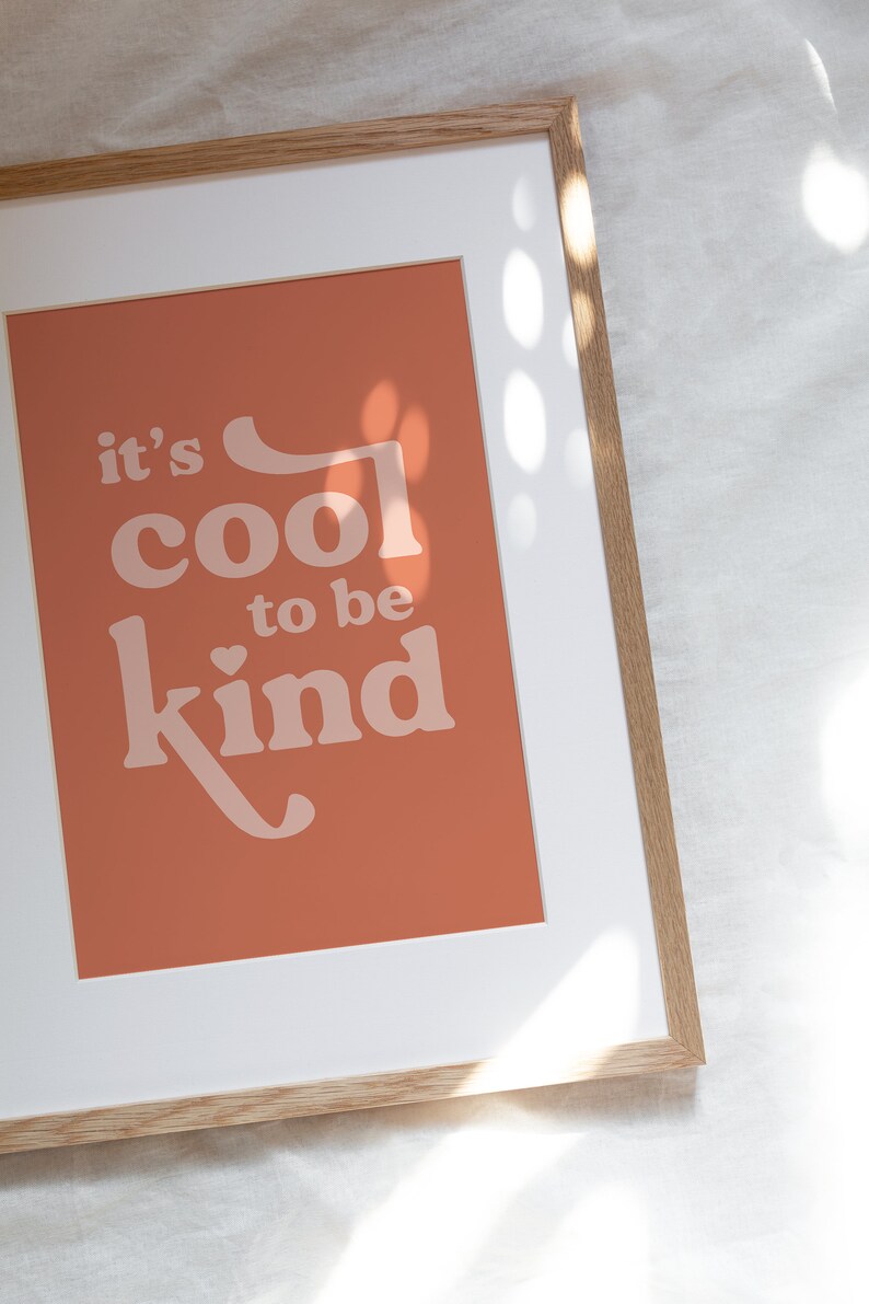 May include: Framed art print with the message "it's cool to be kind" in a retro font. The print has a terracotta background and is framed in a light wood frame with a white mat.