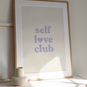 Może przedstawiać: Wydruk w ramie z tekstem "self love club" w jasnofioletowym kolorze. Wydruk znajduje się w jasnej drewnianej ramie i leży na białej powierzchni.