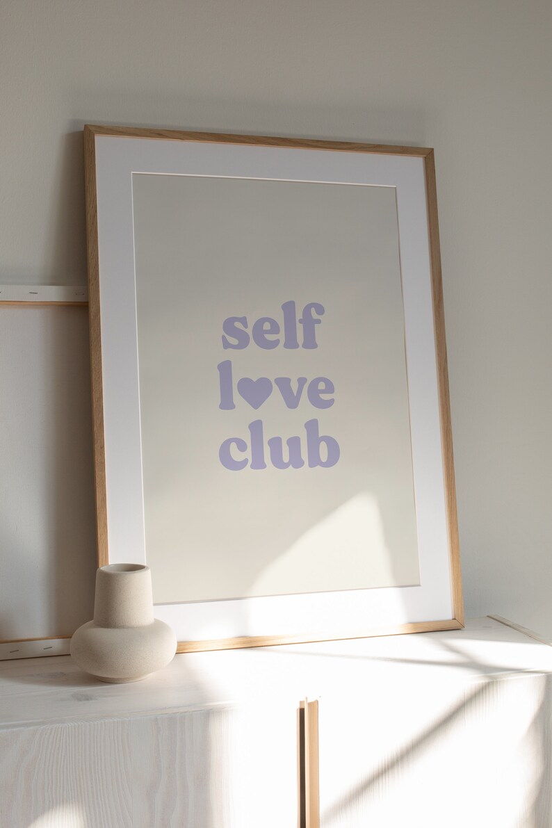 Może przedstawiać: Wydruk w ramie z tekstem "self love club" w jasnofioletowym kolorze. Wydruk znajduje się w jasnej drewnianej ramie i leży na białej powierzchni.