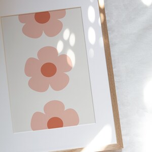 K&ouml;nnte beinhalten: Gerahmter Kunstdruck mit drei stilisierten rosa Blumen mit rostfarbenen Zentren auf cremefarbenem Hintergrund. Das Kunstwerk ist in einem wei&szlig;en Passepartout und einem Naturholzrahmen eingefasst, was eine klare, moderne &Auml;sthetik schafft.
